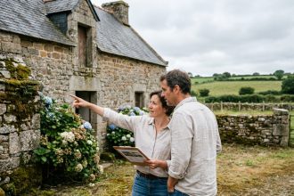 Couple consultant une annonce immobilière devant une maison en pierre bretonne avec terrain et murs en granit